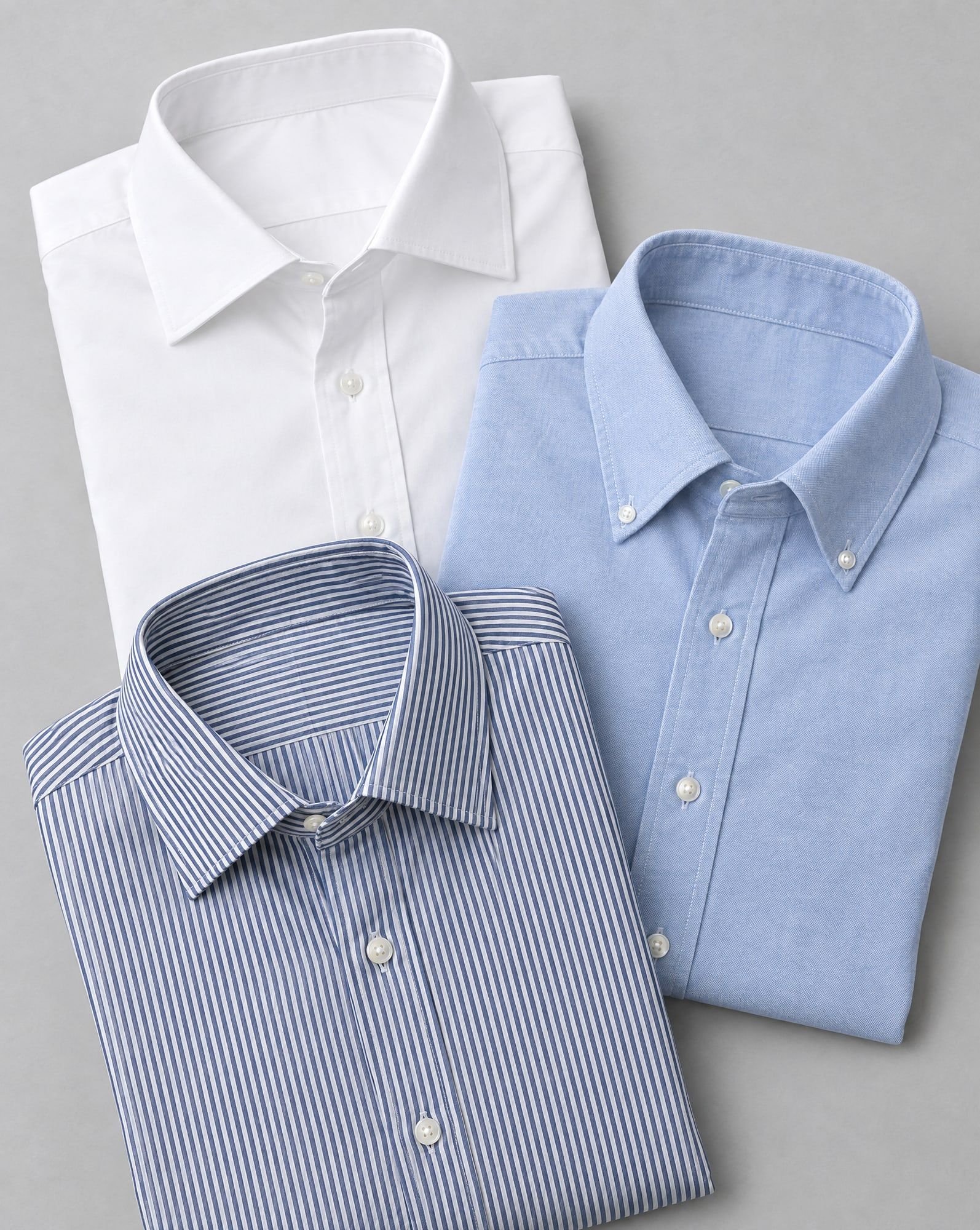 Three folded men's woven cotton shirts in white poplin, pale blue oxford and navy bengal stripe